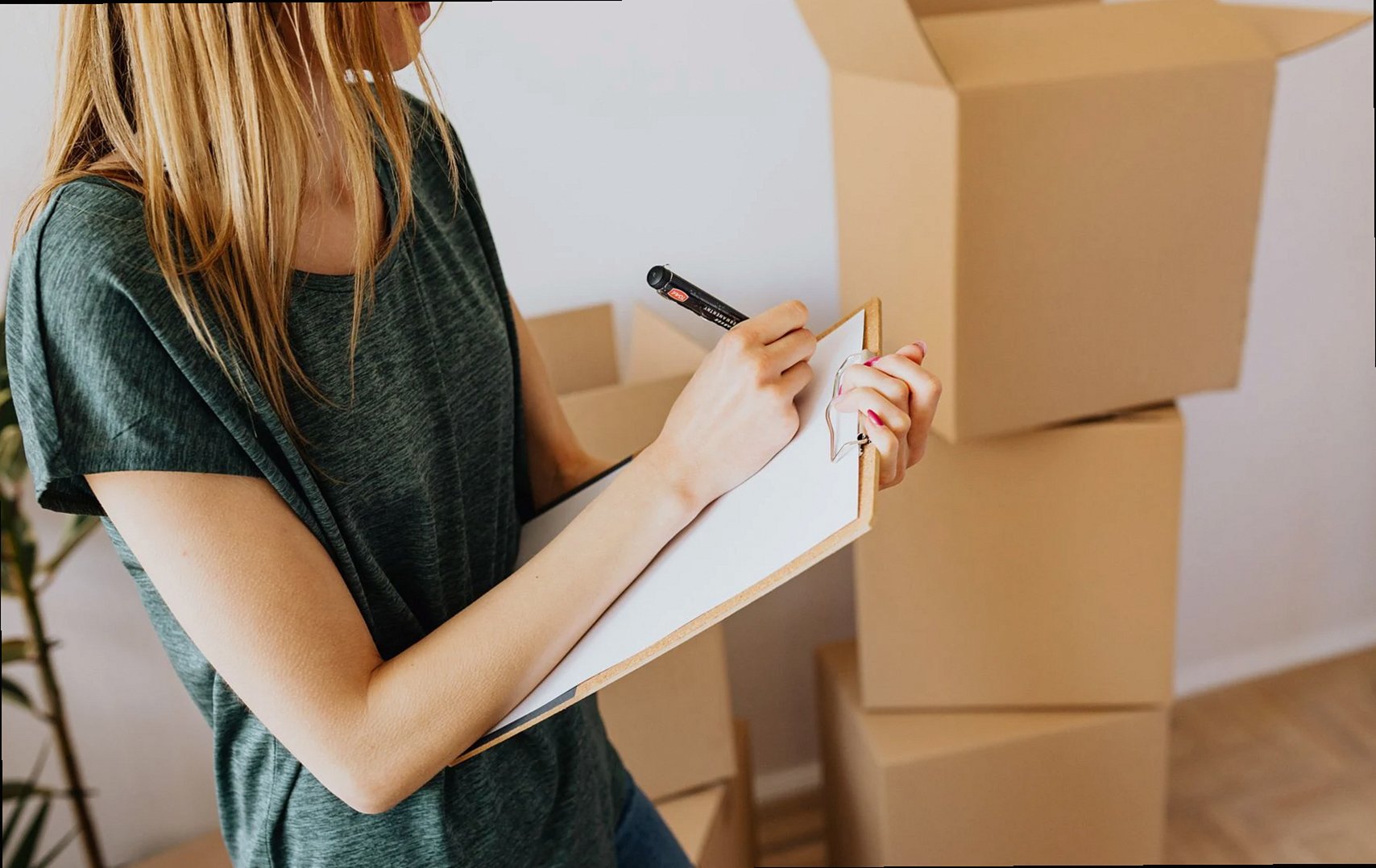 Side view crop female wearing casual shirt standing near packed cardboard containers and taking notes before relocation 